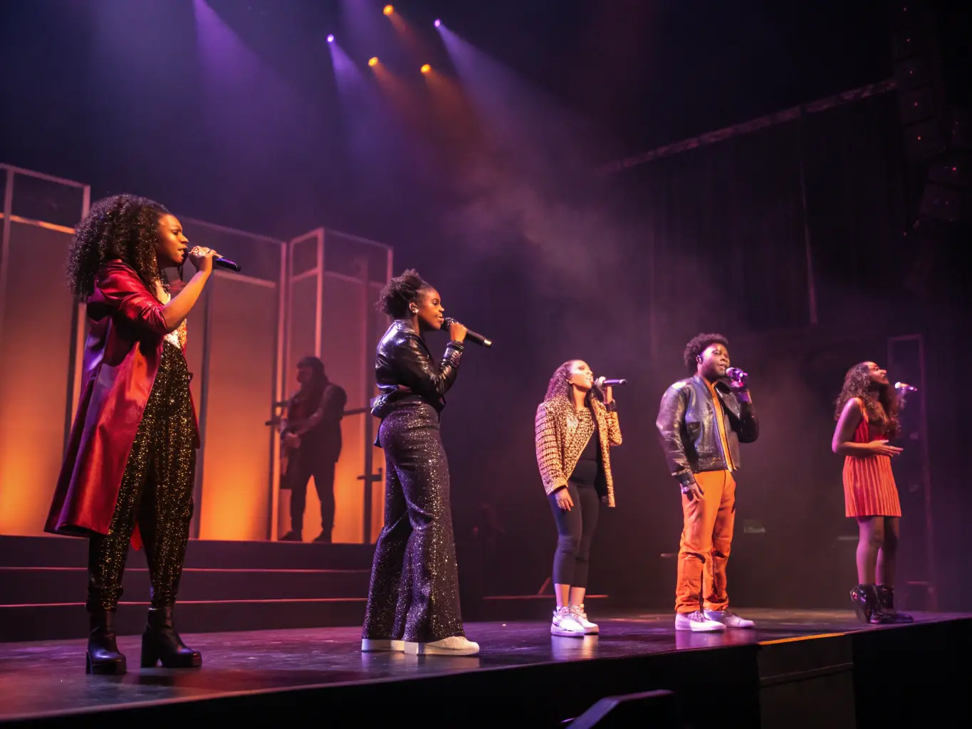 A dynamic image of a live performance by LE PILLI-COLLECTIF, featuring a mix of actors, dancers, and musicians on stage, illuminated by colorful lighting.
